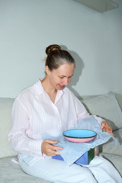 A woman sits on a couch smiling as she unwraps a gift. 