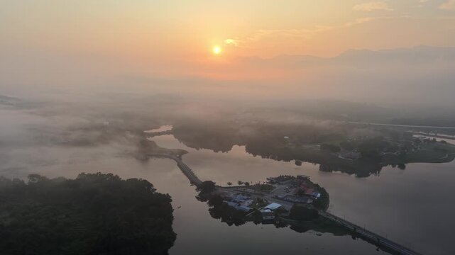 Golden sunrise glows over misty water. Fog blankets islands and calm lake surface at Tasik Raban