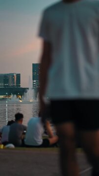 People relax by lake with fountain at sunset in city Benchakitti park skyline, Group sitting by water and enjoy evening view, QSNCC Bangkok Thailand, 4k vertical video