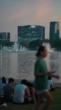 People relax by lake with fountain at sunset in city Benchakitti park skyline, Group sitting by water and enjoy evening view, QSNCC Bangkok Thailand, 4k vertical video