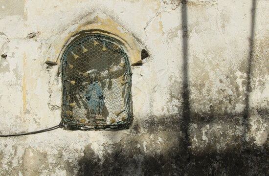 Historische Wandnische mit kleiner Madonnenstatue hinter Drahtgeflecht an einer verwitterten Hauswand