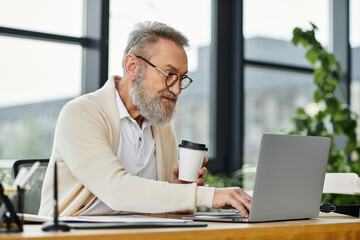 Fototapeta premium Senior man focused on his laptop, sipping coffee during a productive session in the office.
