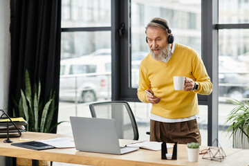 Fototapeta premium Senior man in yellow sweater interacts with a laptop while sipping coffee at his workspace.