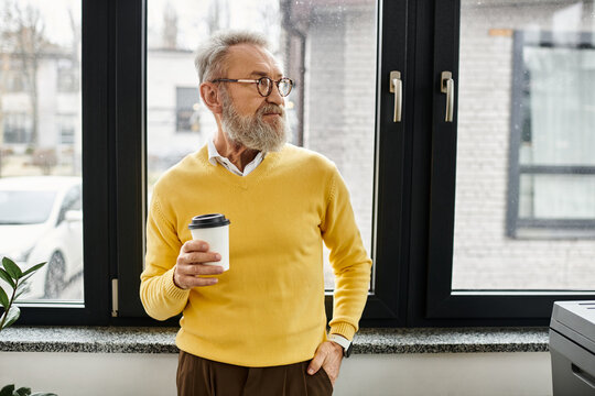 Mature man with a beard holds a coffee cup while gazing thoughtfully out the window.