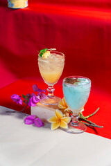Exotic Cocktails on Display: Two vibrant cocktails, one with a pale yellow hue and the other a striking blue, are elegantly presented amidst tropical flowers and garnishes. © andreas