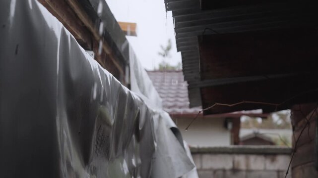 Close-up view of rainwater running and dripping from the edge of a makeshift roof during a daytime downpour. Overcast sky in the background