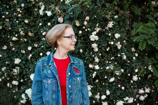 Stylish senior woman 50-55 year old wearing denim jacket and eye glasses over rose flowers outdoors.