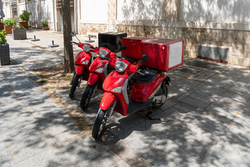 Three red motor scooters equipped with large insulated delivery boxes stand parked together on a paved urban sidewalk. The image represents courier work, restaurant delivery, last mile logistics, take © Volha