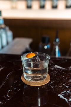 Clear Gin and Tonic on Marble Bar: A refreshing gin and tonic cocktail, garnished with an orange slice and ice, rests on a polished marble countertop in a dimly lit bar setting.