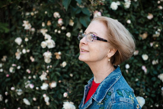 Smiling senior woman 55-60 year old with gray hair and freckles wearing eye glasses over rose flowers outdoors close up. Looking at camera.