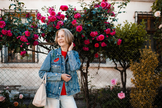 Laughing mature woman 55-60 year old wearing sun glasses and denim jacket outdoors over flowers close up. Looking at camera.