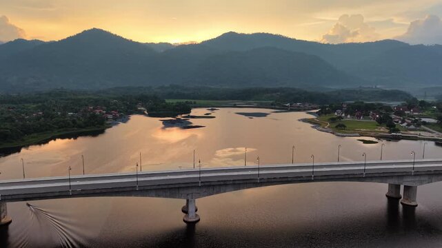 A scenic view of a calm river at sunset with a modern bridge crossing over, surrounded by mountain landscape under a cloudy sky at Tasik Raban