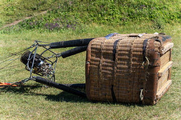 Close-up of a woven hot air balloon gondola with gas burners lying on its side on a green grassy field after landing on a sunny day. © Dina Luk