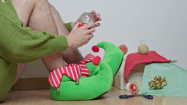 unrecognizable man sitting on the floor wearing elf slippers wrapping christmas presents and shaking a festive snow globe, creating a magical snowfall effect inside the glass ball