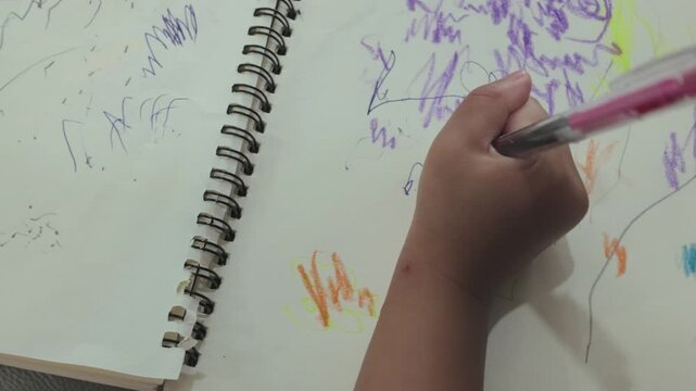 A young child's hand creates vibrant crayon sketches of people and abstract shapes across two pages of a spiral-bound sketchbook.