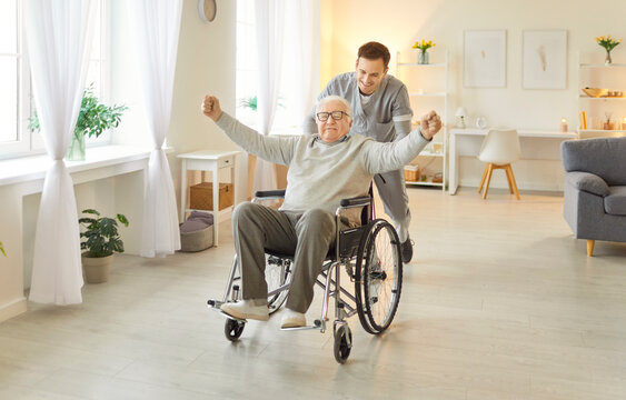 Happy elderly man feels encouraged and motivated clenches fists while posing on wheelchair, being cared by caregiver in nursery house. Male nurse supporting his old patient in rehabilitation