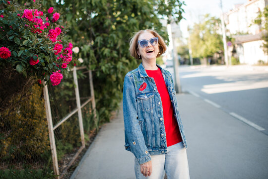 Laughing mature woman 55-60 year old wearing sun glasses and denim jacket outdoors over flowers close up. Looking at camera.