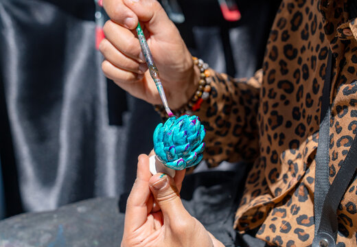 In the foreground, we see the skilled hands of a craftsman busy painting the traditional Sicilian ceramic pine cone &ndash; a symbol of good luck &ndash; in bright colours: turquoise and magenta.