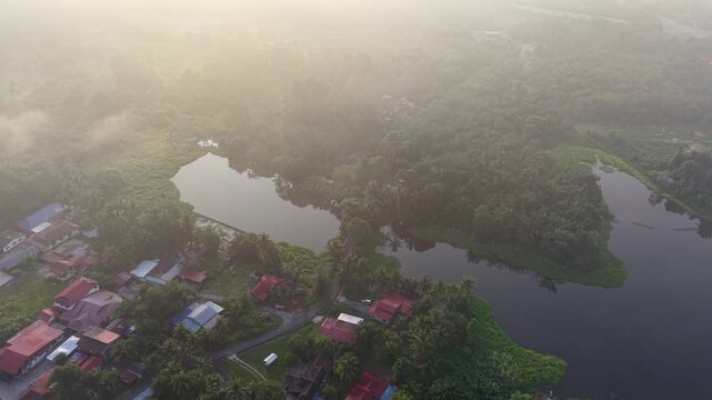 A misty early morning aerial view of a rural village with houses, winding roads, and a river surrounded by dense green forest at Tasik Raban
