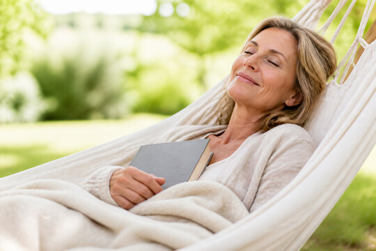 A middle-aged woman is relaxing in a white hammock outdoors, reading a book.