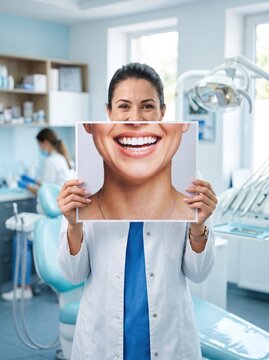Female dentist in white uniform holding photo of perfect white smile over her face inside modern dental clinic office.