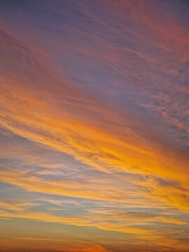 Vibrant sunset sky with multicolor layers of textured clouds. The horizon glows in fiery orange and yellow light, while the upper atmosphere transitions into soft, sweeping lavender and indigo tones