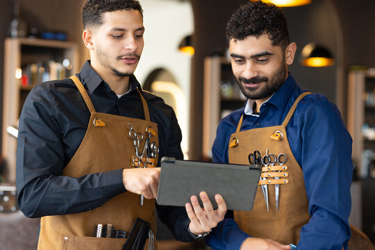 African American male barbers wearing brown leather aprons with scissors, reviewing tablet at salon