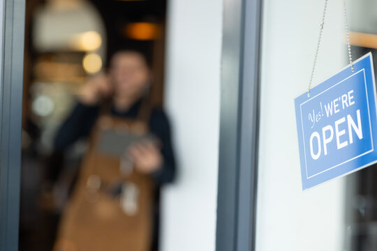 Adult male wearing apron, using smartphone and tablet at shop under blue open sign, copy space