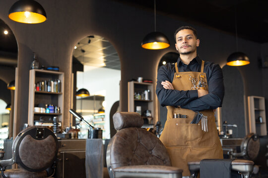 African American barber standing arms crossed chair in shop wearing apron with scissors, copy space