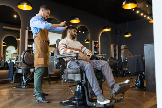 African American male barber with male client at shop, wearing apron cutting hair in classic chair