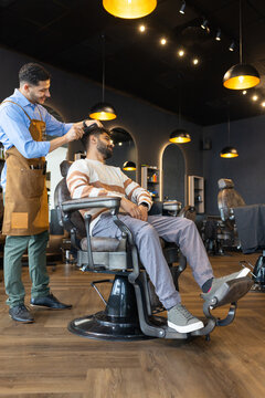 African American male barber and male client at barbershop, wearing apron cutting hair with clipper