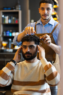 Male barber and client styling short curly hair at salon using comb scissors wearing brown apron