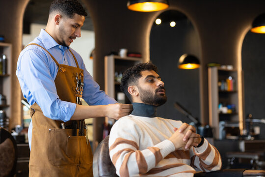 African American male barber securing neck strip, male client sitting in chair at shop, copy space
