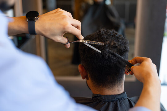 African male barber and client, barber trimming short curly hair using shears and comb at salon