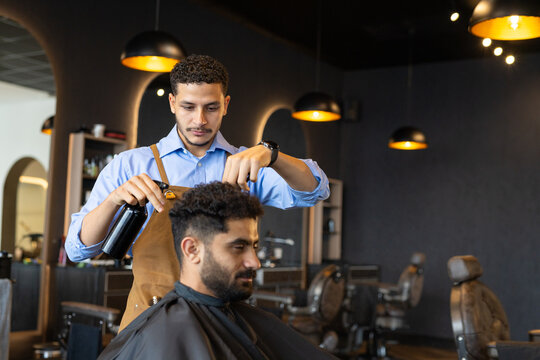 Male barber styling hair for male client in black cape with sprayer and scissors at barbershop
