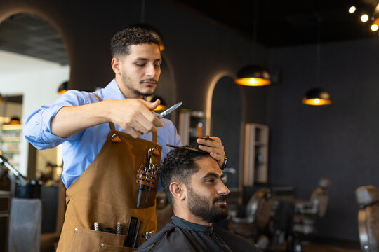 African American men cutting client's hair in barbershop with scissors and brown apron
