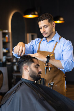 African male barber trimming male client hair with scissors and comb in leather apron at barbershop
