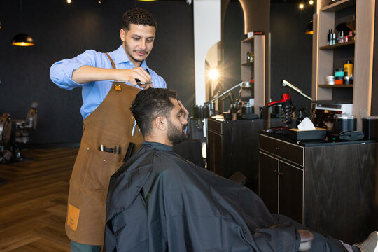 Male barber and male client cutting hair at barbershop station, wearing apron and using scissors