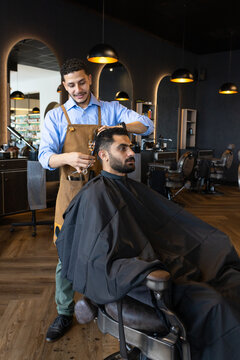 African American barber and client trimming hair with scissors, apron and black cape at shop