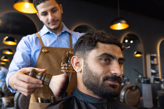 Male barber brushing client neckline using neck brush in barbershop, client wearing black cape