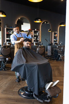 African American males, barber cutting client's hair using hair dryer and comb in shop, brown apron