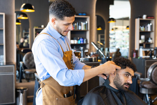 Two African men in barbershop, barber trimming client hair with trimmer, brown apron and black cape