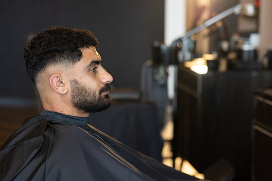 Adult man sitting in barber chair at barbershop wearing black cutting cape, copy space