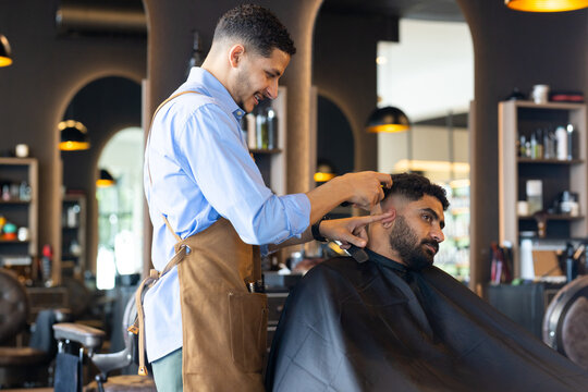 African American barber trimming client's hair with clippers, steadying ear at shop, wearing apron
