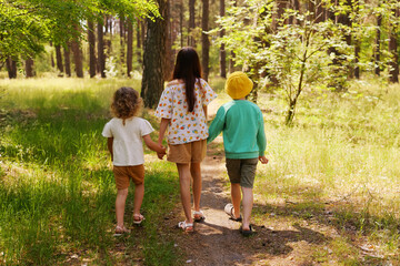 Fototapeta premium Three children stroll down a dirt path in a forest. They hold hands and enjoy the sunny day surrounded by trees and plants