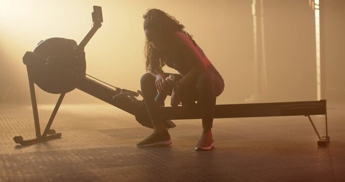 After rowing interval African American woman sitting on erg in gym holding water bottle recovering