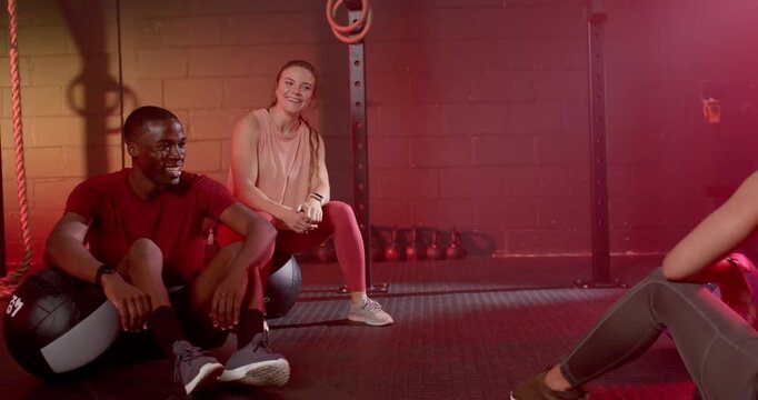 Diverse friends laughing and gesturing after off-frame coach cue, resting on exercise balls in gym