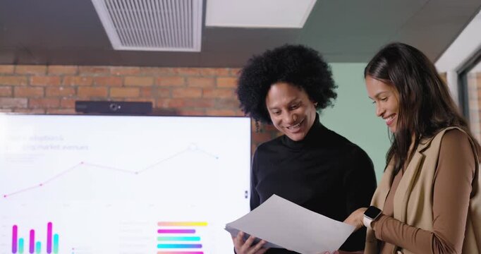 Two African American women being prompted by screen charts at office reviewing tablet and papers