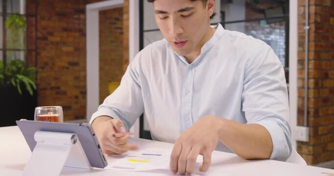 Adult Asian man glancing between tablet and printed charts, annotating reports with pen at desk