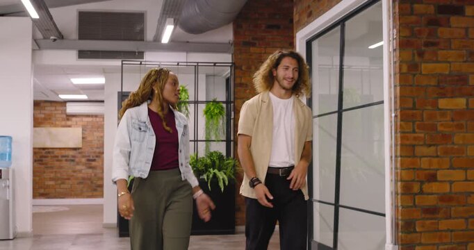 Diverse coworkers walking in corridor, man gesturing, woman sitting on pink ottoman continuing chat